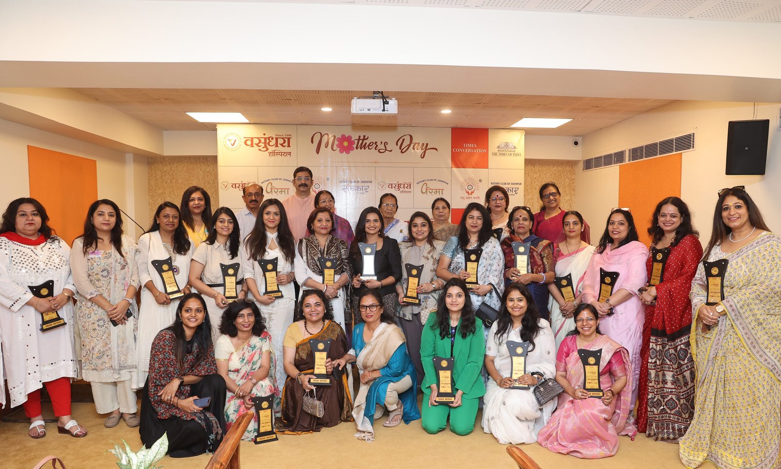 Group of women in colorful traditional attire posing on stage, each holding a trophy, with a Mother's Day banner in the background.